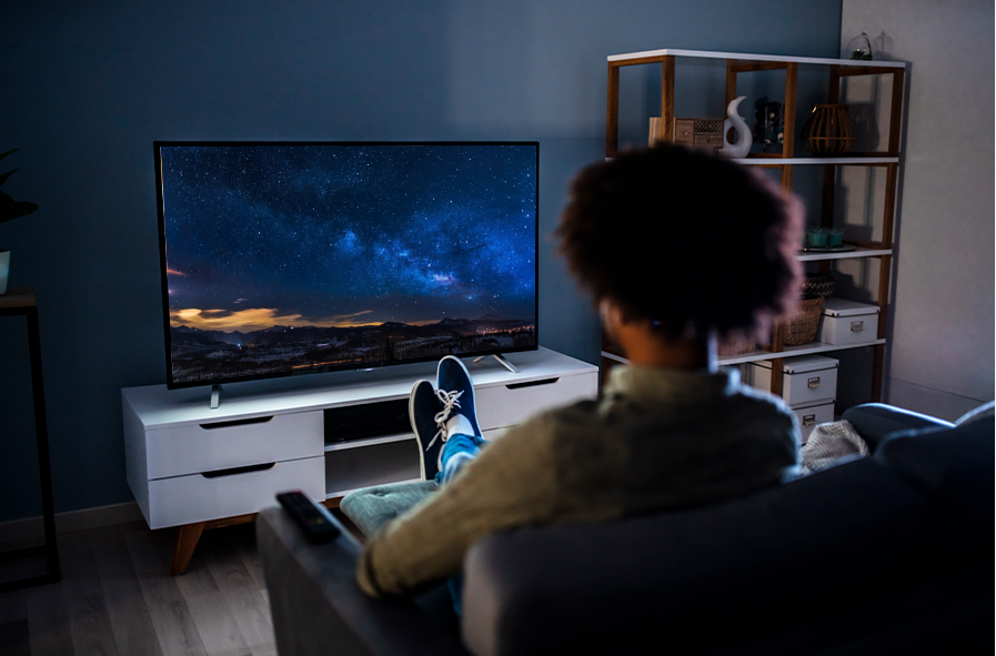 Man sitting on a grey couch with his back to foreground and his feet up on an ottoman, watching TV in modern living room